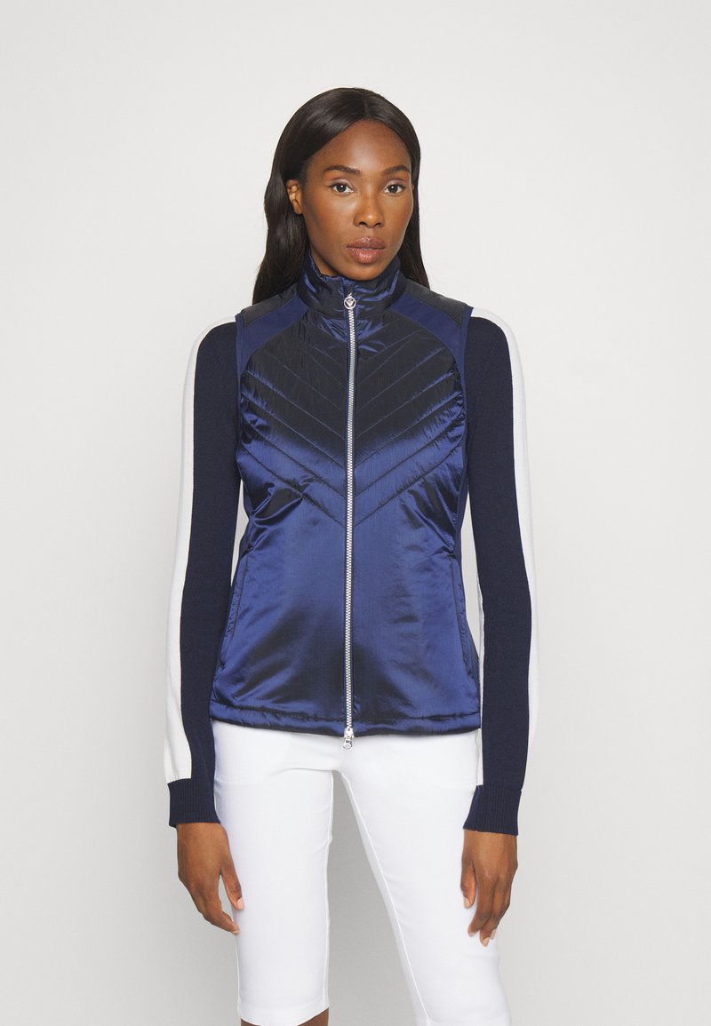 Modrá vycpaná vesta s vysokým límcem, zipem vpředu a kontrastními tmavými rukávy. Má design prošívání ve tvaru češky a hladký povrch.