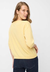 Pull en maille jaune avec manches trois-quarts, coupe décontractée et encolure arrondie, associé à un jean foncé. Texture douce.