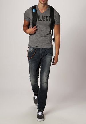 T-shirt grigio a scollo a V con la parola "EJECT" stampata, etichetta blu. Jeans in denim blu con scoloriture, catena accessoria marrone e sneakers grigie. Zaino nero.