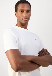 Witte t-shirt met korte mouwen, ronde hals en kleine merktekst op de borst. Glad stof, losse pasvorm en lichte textuur.