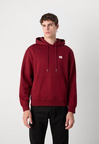 Felpa bordeaux in cotone con tasca a canguro, coulisse regolabile e patch con logo sul petto. Polsini e orlo ribattuti.