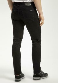 Czarne jeansy slim fit z denimu, z tylnymi kieszeniami z poziomowym zamkiem błyskawicznym i subtelnymi detalami przeszycia. Noszone z czarnymi trampkami.