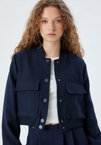 Marineblaue cropped Jacke aus strukturiertem Stoff, mit zwei Brusttaschen, Druckknöpfen sowie gerippten Bündchen und Kragen.