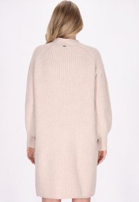 Vrouw met lang blond haar in een beige, grof gebreid sweaterjurk, gezien van achteren tegen een eenvoudige witte achtergrond.