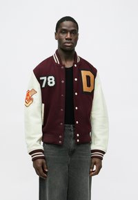 Tamsiai raudonos ir kreminės spalvų universiteto stiliaus striukė su "D" raidės patch'u, ant krūtinės išsiuvinėtas skaičius 78 ir dryžuoti megzti rankogaliai. Audinys yra lygios tekstūros.