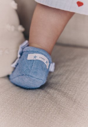 Dziecko noszące niebieski materiałowy but z etykietą „happy” na pięcie, siedzące na teksturowanej jasnoszarej powierzchni.