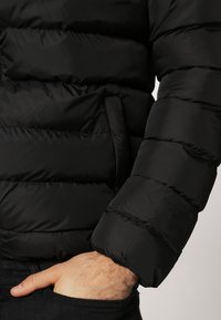 Chaqueta de plumas negra con secciones acolchadas, una textura suave y puños ajustados. El material parece ligero pero aislante.