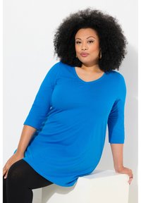 Electric blue, strak zittend langslurf shirt met een V-hals en halflange mouwen, gecombineerd met zwarte leggings, zittend tegen een lichte achtergrond.