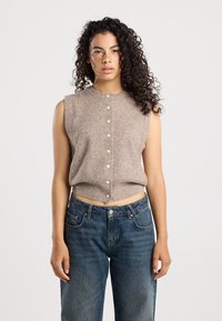 Débardeur tricoté cropped, sans manches, de couleur marron clair, avec une fermeture à boutons sur le devant. Porté avec un jean en denim bleu foncé, taille haute.