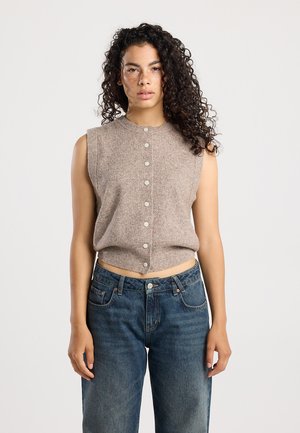 Débardeur tricoté cropped, sans manches, de couleur marron clair, avec une fermeture à boutons sur le devant. Porté avec un jean en denim bleu foncé, taille haute.