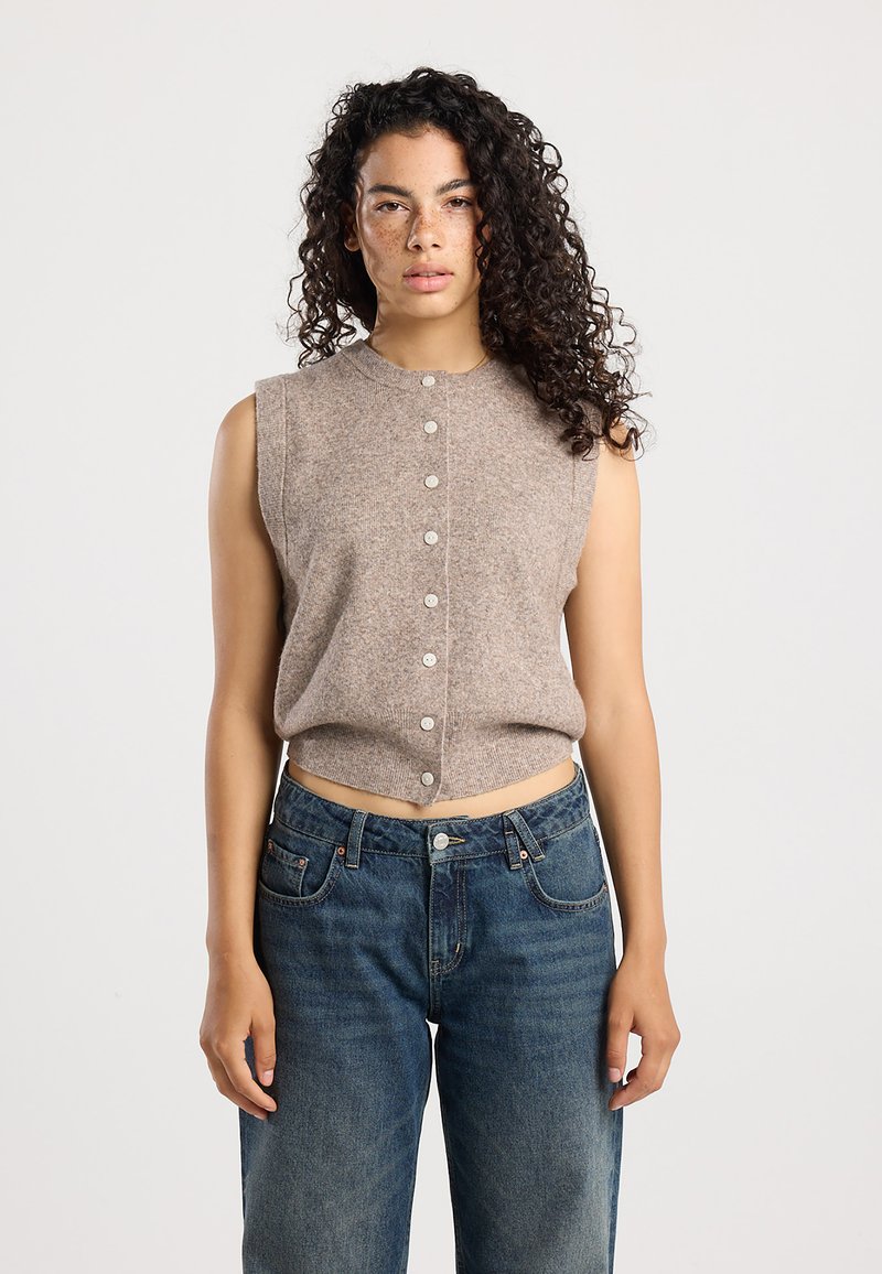 Débardeur tricoté cropped, sans manches, de couleur marron clair, avec une fermeture à boutons sur le devant. Porté avec un jean en denim bleu foncé, taille haute.