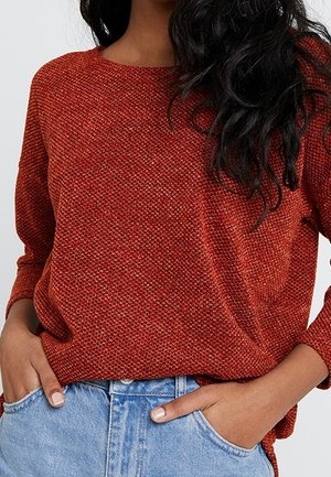 Femme portant un pull en maille texturée de couleur rouille à manches longues, rentré dans un jean denim bleu clair, les mains dans les poches.