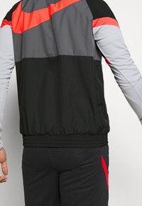 Chaqueta de pista negra y gris con acentos rojos, que cuenta con cierre de cremallera, puños elásticos y un diseño de tela texturizada.