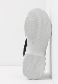 Suola in gomma bianca di una sneaker nera che mostra il motivo del battistrada, inclinata verticalmente contro una superficie angolare bianca.