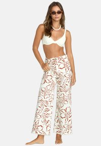 Weiße, weite Blumen-Hosen mit braunem Blumenmuster, kombiniert mit einem cremefarbenen Oberteil. Der Stoff wirkt weich, das Design ist leger.