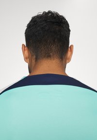Camiseta deportiva turquesa con cuello azul marino, con textura suave y mangas cortas, vista desde la espalda.