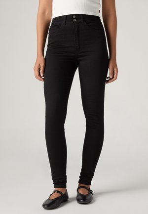 Femme portant un jean skinny noir taille haute, un haut blanc sans manches et des chaussures Mary Jane noires, debout devant un fond uni.