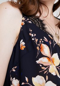 Blommig linne med marinblå bakgrund, prydd med orange och vita blommor. Spetsdetalj längs u-ringning och mjukt tyg. U-ringningen är i v-form.