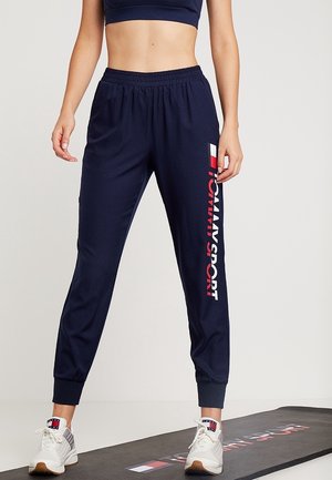 Kobieta ubrana w granatowe spodnie dresowe Tommy Sport i białe trampki stoi na czarnej macie do ćwiczeń z logo Tommy Sport.