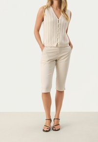 Beige gebreide mouwloze cardigan met V-hals en knopen, gecombineerd met lichtbeige knielange broeken en zwarte sandaaltjes met bandjes.