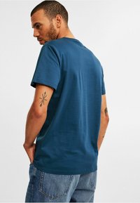 Mand, der står med ryggen til, iført en blå T-shirt med korte ærmer og blå jeans, der viser tatoveringer på begge arme foran en ensfarvet baggrund.