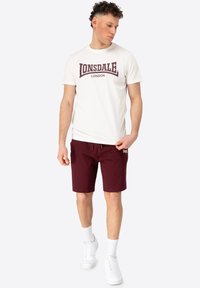 Hvid bomulds T-shirt med maroon "LONSDALE LONDON" logo, parret med maroon shorts. Modellen bærer hvide sneakers og strømper i crew-længde.