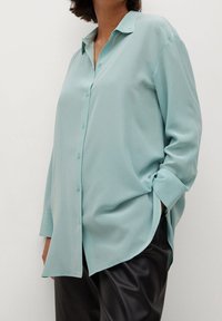 Chemise bleu clair à manches longues, boutonnée, en tissu léger avec une coupe décontractée et un léger éclat. Présente un col pointu et des fentes latérales.