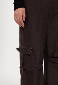 Pantalons cargo marron à rayures avec une poche inclinée, des coutures orange contrastantes et une coupe décontractée. Le matériau semble léger et texturé.