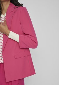 Fuchsia blazer med skräddarsydd design, enknappsstängning och ficka. Lager på en vit texturerad topp med rosa Accenter.