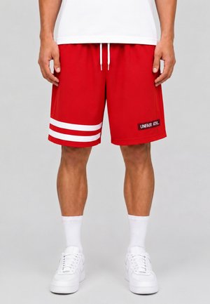 Mand iført røde atletikshorts med hvide striber på venstre ben og "UNFAIR ATHL" plet på højre, hvide sokker og hvide sneakers.