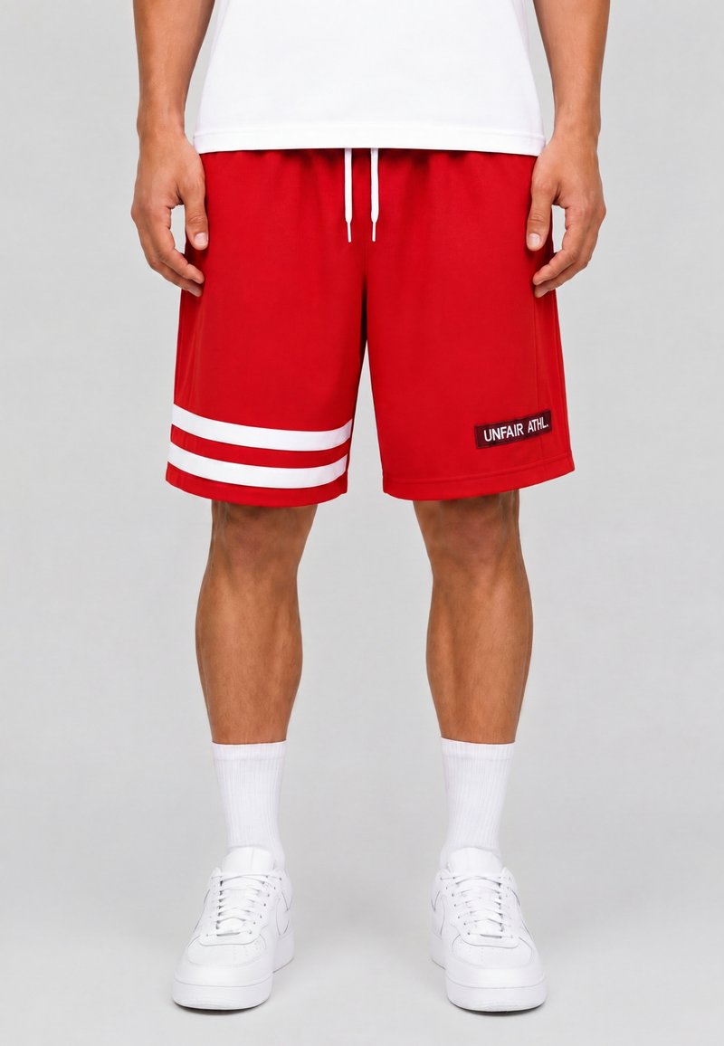 Homme portant un short de sport rouge avec des rayures blanches sur la jambe gauche et un patch "UNFAIR ATHL" à droite, chaussettes blanches et baskets blanches.