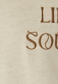 Romige gebreide stof met daarop een bruine geborduurde tekst die "LIFE" en "SOUL" spelt. De textuur is glad met een losse, casual uitstraling.
