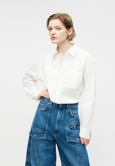 Νεαρός με κοντά μαλλιά που φοράει λευκό πουκάμισο με κουμπιά και μπλε ψηλόμεσα τζιν παντελόνια cargo, στέκεται με το ένα χέρι στη μέση.