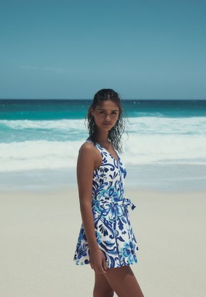 Femme aux cheveux mouillés portant une robe fleurie bleue et blanche, debout sur une plage de sable avec des vagues de l'océan en arrière-plan.