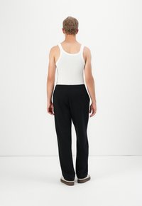 Persona de pie de espaldas, vistiendo una camiseta sin mangas blanca, pantalones negros y zapatillas blancas, frente a un fondo blanco liso.