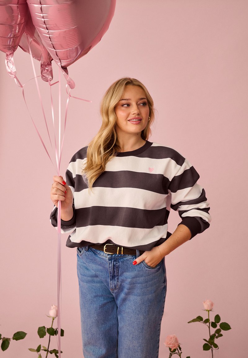 Mujer con cabello largo y rubio sostiene globos rosados en forma de corazón, vestida con suéter de rayas negras y blancas y jeans azules, de pie junto a rosas rosas.