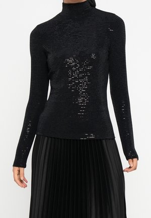 Schwarzes, langärmliges, mit Pailletten besetztes Rollkragenoberteil kombiniert mit einem hoch taillierten, schwarzen, plissierten Rock an einer stehenden Person.