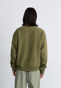 Oseba z dolgimi temnimi lasmi se obrne stran, oblečena v olivno zeleno majico in svetlo zelen hlače, pred enobarvno belo ozadje.