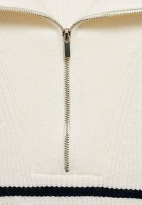 Stickad halvdraget tröja i krämfärgad med ribbad textur, med en silverblixtlås och ett svart horisontellt rand nära fållen.