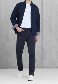 Námořnická košile na knoflíky s vyhrnutými rukávy přes bílé tričko, doplněná tmavými kalhotami a bílými teniskami, na pozadí z betonu.