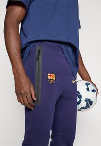 Paarse sportbroeken met een zijzak met rits, geborduurd Barcelona-logo en een bijpassend blauw shirt. Een voetbal wordt in één hand vastgehouden.