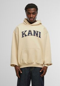 Beige oversized hoodie med en framficka, med mörkblå broderad text "KANI". Slät textur; långa ärmar med ribbade muddarna.