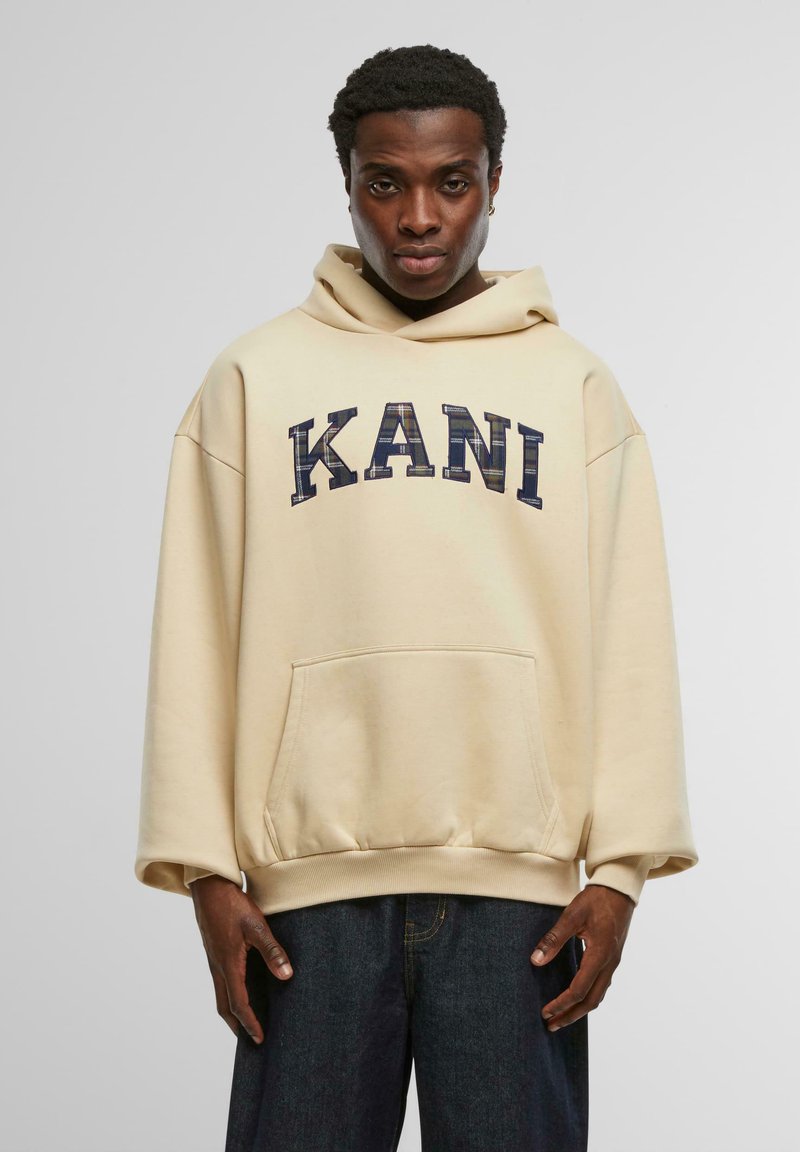 Beige oversized hoodie med en framficka, med mörkblå broderad text "KANI". Slät textur; långa ärmar med ribbade muddarna.