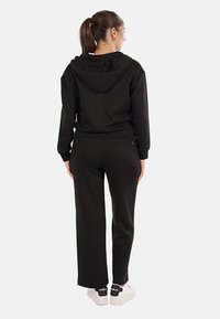 Felpa nera con cappuccio e pantaloni abbinati realizzati in tessuto morbido. I pantaloni hanno una vestibilità comoda. Sneakers bianche visibili, con marchio sul tallone.