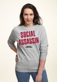 Grå crewneck-tröja med fet röd text "SOCIAL ASSASSIN" och en mindre svart "Curb Your Enthusiasm"-logotyp. Slät textur, avslappnad passform.