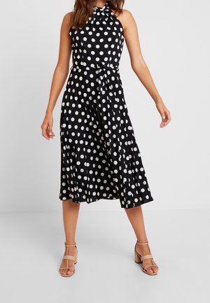 Robe noire sans manches à pois blancs, jupe plissée et ceinture nouée à la taille. Le tissu semble léger, avec une texture lisse.