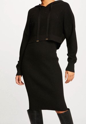 Frau trägt ein schwarzes, geripptes Midikleid mit langen Ärmeln und einem passenden kurzen Hoodie, kombiniert mit schwarzen kniehohen Stiefeln.