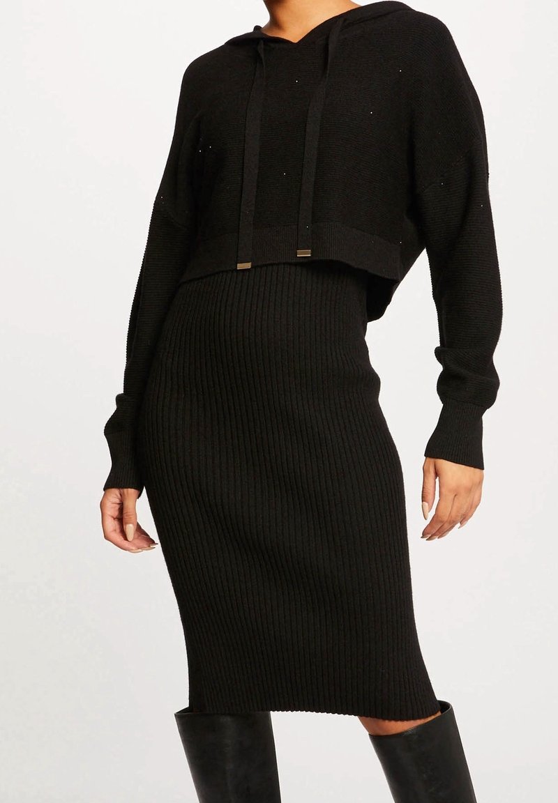 Femme portant une robe midi en tricot côtelé noire à manches longues et un sweat à capuche court assorti, associée à des bottes noires montant jusqu'aux genoux.