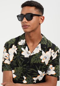Cămașă florală neagră cu mâneci scurte, decorată cu flori albe și portocalii, frunze verzi și ochelari de soare negri purtată de un tânăr. Textura materialului este fină.