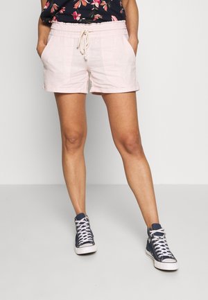 Person iført lyserøde snøre-shorts, navyblå blomstret top og navy høje sneakers stående med hænderne i lommerne.